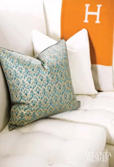 White tufted sofa with a teal and gold patterned throw pillow and an orange throw blanket with a white H logo.