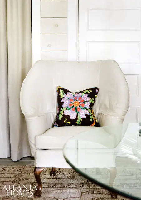 Cream upholstered armchair with a dark throw pillow featuring a colorful floral pattern, set against a white paneled wall and neutral curtains.