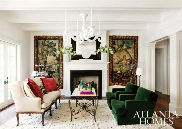 Living room with a cream sofa, two emerald green armchairs, a fireplace flanked by tapestries, and a white chandelier.