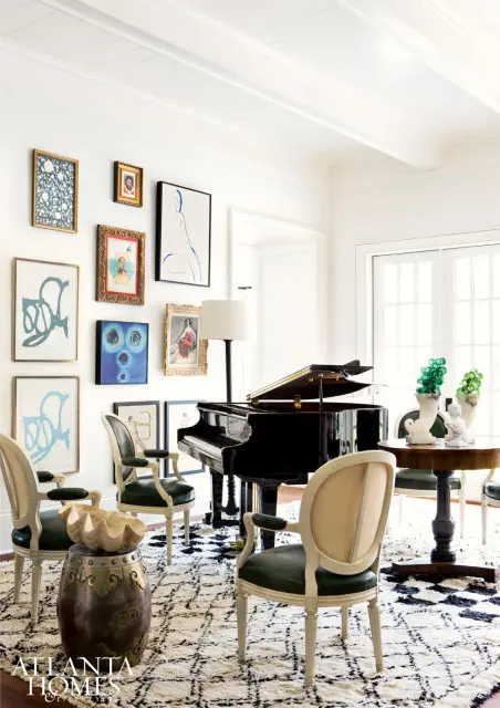 Living room with a grand piano, dark green upholstered chairs, a round wooden table with decorative vases, and a collection of framed art on a white paneled wall.