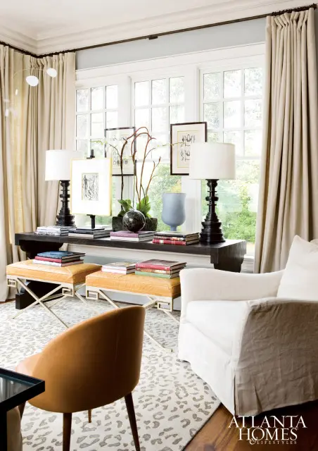 Living room with a dark wood console table displaying lamps, books, and decorative objects in front of a large window with beige curtains. Two orange upholstered ottomans sit on a leopard print rug.
