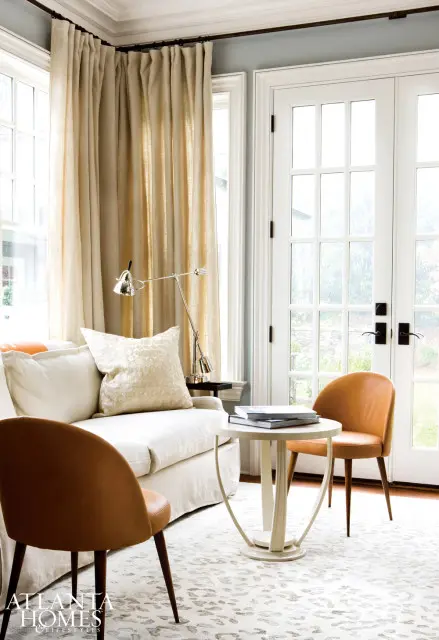 Living room with a cream slipcovered sofa, two tan leather accent chairs, and a white round side table with books. Large beige curtains hang over a window, and French doors lead outside.