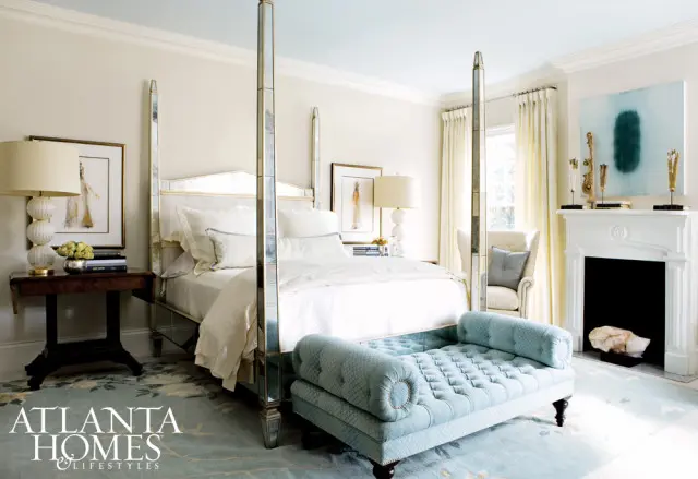 Bedroom with a mirrored four-poster bed, tufted light blue chaise lounge, white fireplace, and a light blue rug with floral accents.