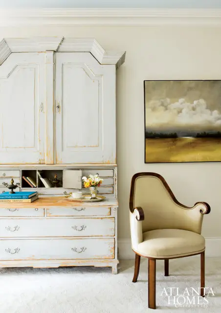Light blue distressed secretary desk with gold accents, a cream upholstered chair with dark wood trim, and a landscape painting on a cream wall.