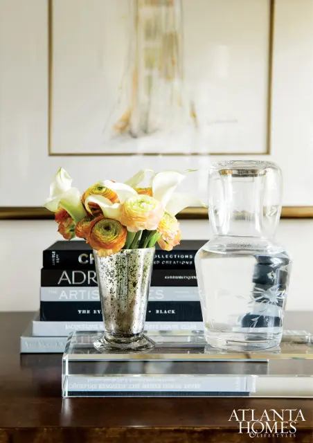 A bouquet of white calla lilies and orange ranunculus in a speckled silver vase sits on a stack of books next to an etched glass carafe on a dark wood table.