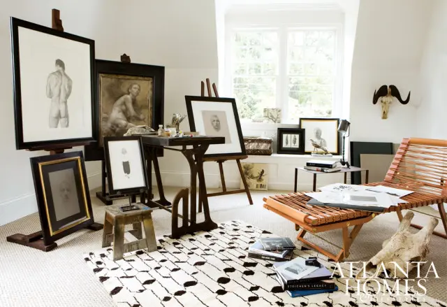 Artist studio with framed artwork on easels, a wooden desk with art supplies, a leather lounge chair, and a patterned rug.