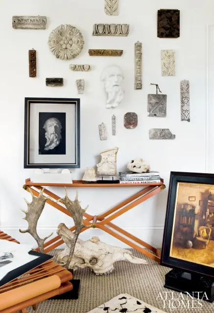 Wall display of antique architectural fragments and sculptures, including a bust of a man, above a console table with antlers, a skull, and books.