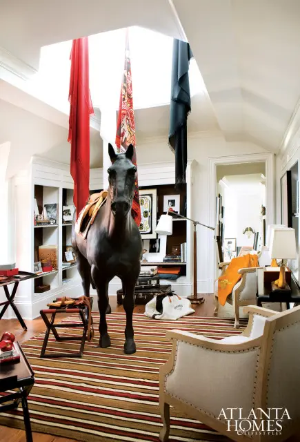 Interior room with a dark horse statue wearing a saddle, red, patterned, and blue scarves hanging from the ceiling, built-in bookshelves, striped rug, and upholstered chairs.