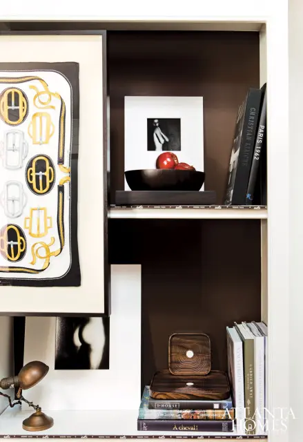 White bookshelf with dark brown backing displaying framed art, books, and decorative objects including a wooden bowl with apples and a vintage desk lamp.