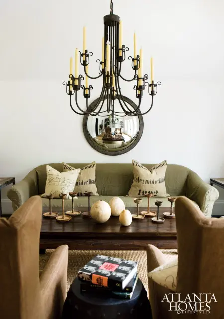Olive green sofa with burlap pillows and a dark wood coffee table holding decorative spheres and candle holders, beneath a wrought iron chandelier and round mirror.