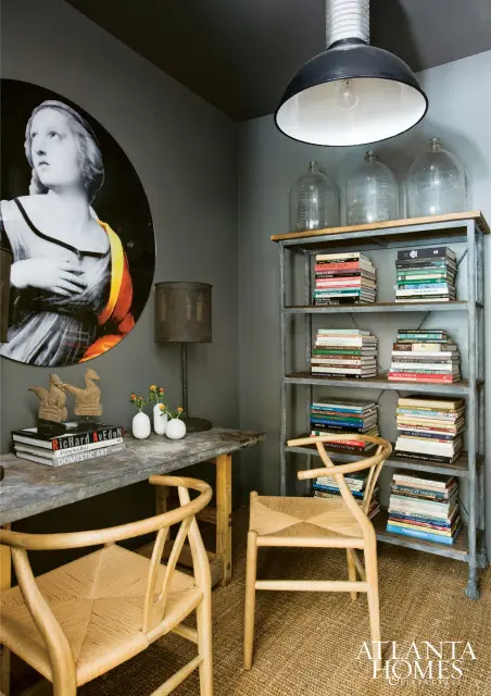 Dark gray room with a large circular artwork of a woman, a metal desk with books and vases, two woven chairs, and a metal bookshelf filled with books.