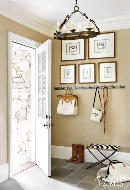 Entryway with a chandelier, framed equestrian prints, a coat rack with two handbags, brown boots, a zebra-print stool, and a stone wall visible through an open door.