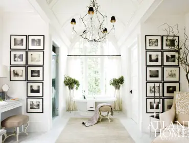 Bright white bathroom with a freestanding tub, arched window, crystal chandelier, vanity, and gallery walls of black and white photographs.
