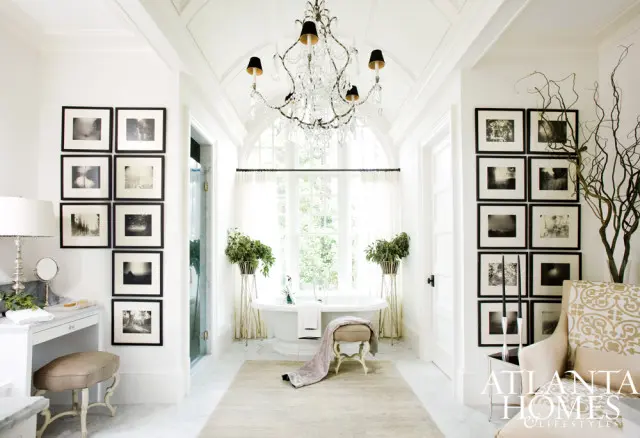 Bright white bathroom with a freestanding tub, arched window, crystal chandelier, vanity, and gallery walls of black and white photographs.