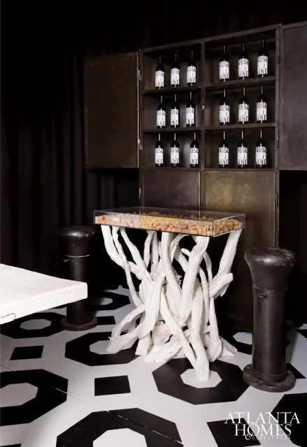 Dark metal cabinet filled with bottles of wine, a white sculptural console table with a glass top filled with corks, and a black and white geometric patterned floor.