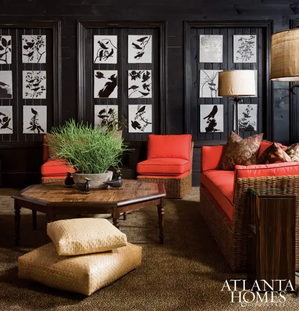 Living room with black paneled walls adorned with framed bird art, featuring coral upholstered wicker furniture, a hexagonal bamboo coffee table, and leopard print carpet.