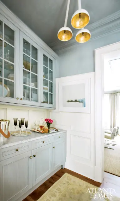 Light blue kitchen cabinets with glass fronts, marble countertops, and brass hardware. Above, a gray ceiling with a cluster of gold pendant lights.