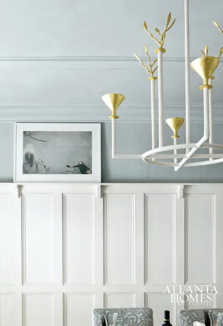 Light blue textured wallpaper above white paneled wainscoting, with a framed artwork and a white chandelier with gold accents.