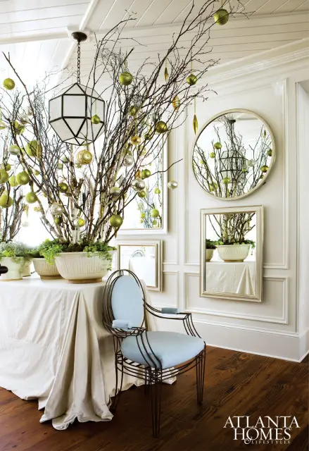 Dining room with a table draped in a white cloth, adorned with bare branches decorated with green and gold ornaments. A light blue upholstered metal chair sits nearby, with paneled walls and mirrors reflecting the festive display.
