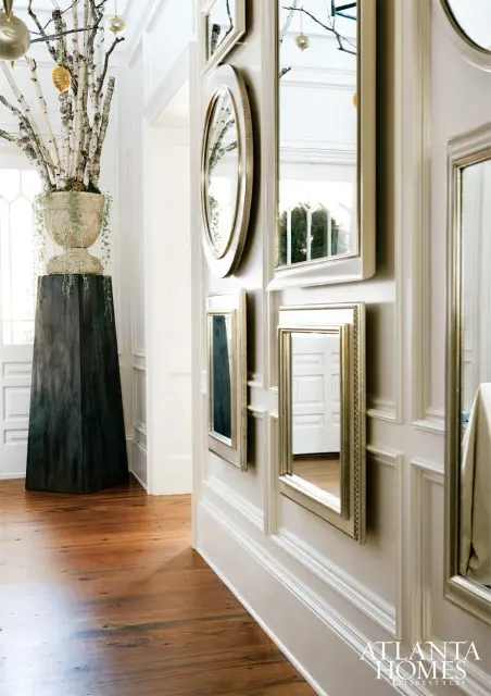 Hallway with paneled walls adorned with a collection of gold-framed mirrors, a large urn with birch branches, and dark wood floors.