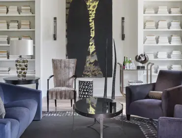 Living room with blue sofa, upholstered armchairs, black coffee table, and built-in bookshelves filled with books. A large abstract painting hangs on the wall.
