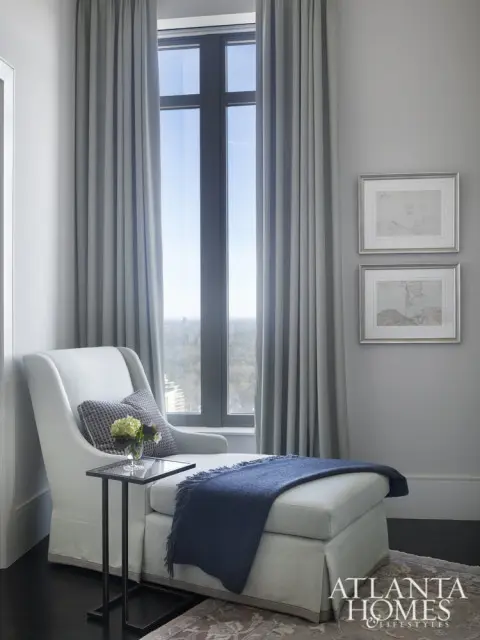 Light blue chaise lounge with a navy blue throw blanket and decorative pillows next to a tall window with grey curtains. Two framed maps hang on the wall.
