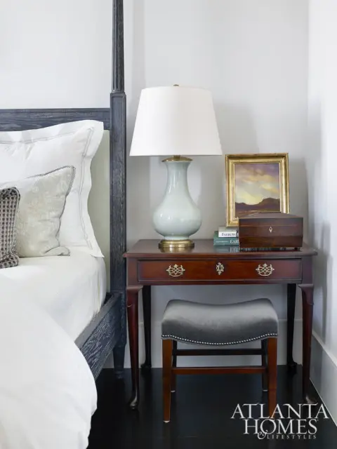 Bedroom with a dark wood four-poster bed, white bedding, a pale blue ceramic lamp on a mahogany side table, and a gray velvet stool.