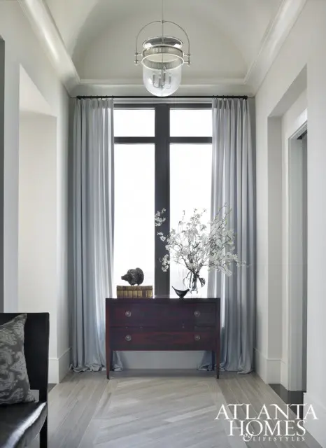 Hallway with arched ceiling, featuring a dark wood dresser beneath a tall window draped with light blue curtains. A glass pendant light hangs overhead, and the floor is tiled in a light wood herringbone pattern.