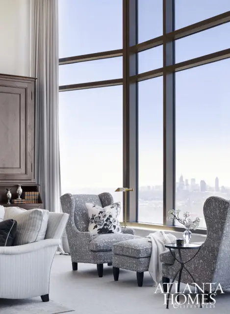 Living room with patterned armchairs and ottomans facing a large window with a city view. A dark wood cabinet is on the left.