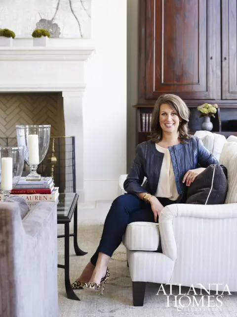 Woman in a navy jacket and jeans sitting in a striped armchair in a living room with a fireplace and large wooden cabinet.