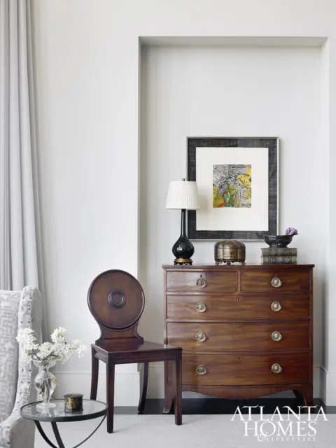 Living room vignette with a mahogany chest of drawers, a dark wood chair with a circular back, and a framed artwork above a lamp.
