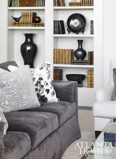 Dark gray velvet sofa with patterned pillows in front of a white built-in bookshelf filled with books and decorative vases. Atlanta Homes & Lifestyles logo in the corner.
