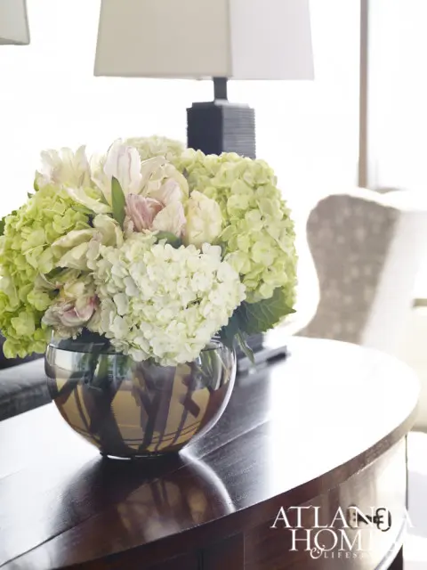 A round glass vase filled with hydrangeas and tulips sits on a dark wood table next to a lamp.
