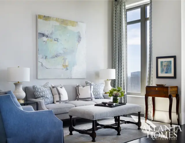Light gray sofa with blue and white patterned pillows, a gray upholstered ottoman, and a blue armchair in a living room with abstract art and a large window.