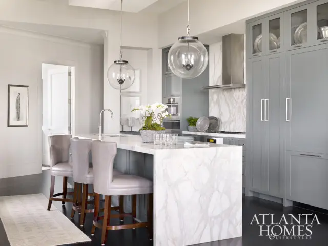 Gray kitchen with marble island, three gray leather bar stools, and gray custom cabinetry with glass-front upper cabinets.