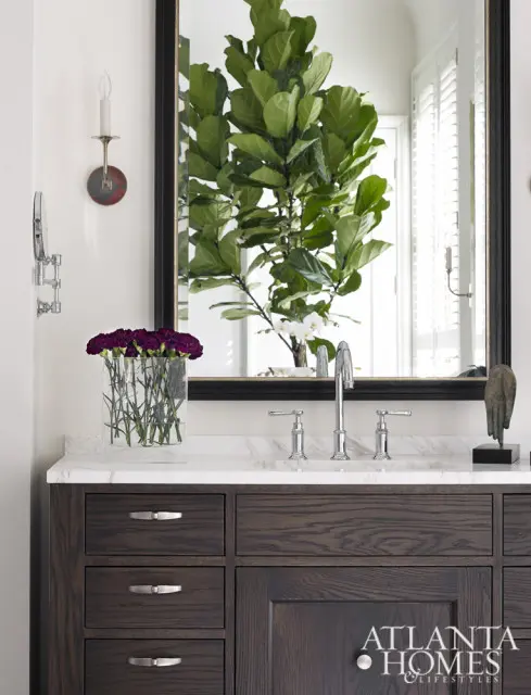 Dark wood vanity with marble countertop and chrome fixtures, reflected in a mirror with a large fiddle-leaf fig tree.