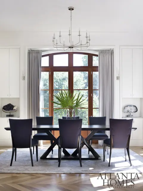 Dark wood dining table with upholstered chairs in front of a large arched window with wood trim. A modern chandelier hangs above.