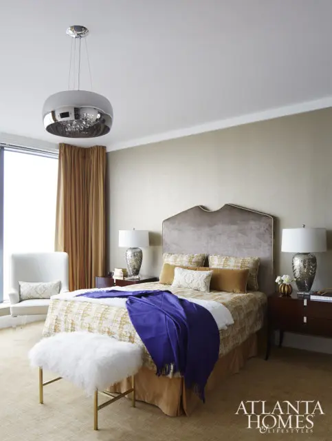 Bedroom with a king-size bed, velvet headboard, patterned bedding, purple throw blanket, white fur bench, and gold curtains.