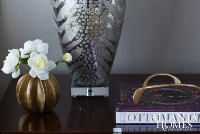 Close-up of a gold fluted vase with white peonies, a large etched glass vase, and a book titled "Ottoman Homes & Lifestyles" on a dark wood surface.