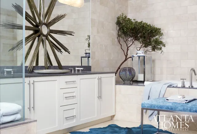 White bathroom vanity with gray countertop, brushed nickel hardware, and a large sunburst mirror above. A blue bench sits next to a bathtub with a light blue towel.
