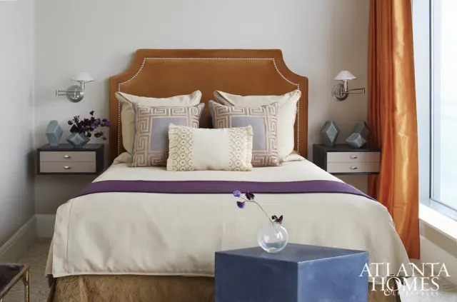 Bedroom with a burnt orange velvet headboard, neutral bedding, purple runner, and decorative pillows. Orange curtains hang by a window.