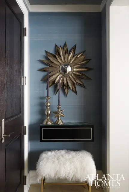 Entryway with dark wood door, blue grasscloth wallpaper, sunburst mirror, brass candlesticks, black floating shelf, and white fluffy bench.