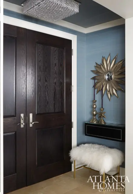 Dark wood double doors with brushed nickel hardware, adjacent to a blue grasscloth wallpapered wall with a sunburst mirror and gold candleholders.