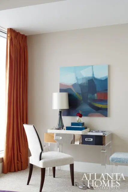 Neutral toned room with a modern desk featuring a clear acrylic frame and drawers with varied textures. A white upholstered chair sits in front of the desk, with a colorful abstract painting and a lamp above.