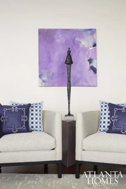 Two cream upholstered armchairs with dark wood frames flank a dark metal sculpture on a pedestal, set against a wall with a large abstract purple painting.