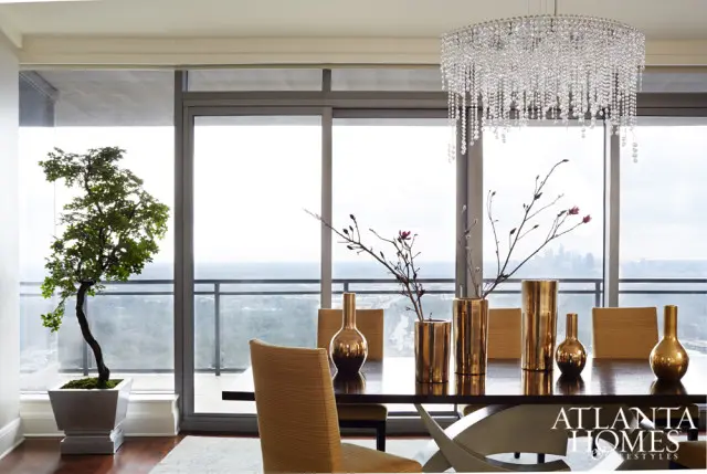 Dining room with large windows overlooking a city skyline, featuring a dark wood table with metallic vases and branches, a crystal chandelier, and a potted bonsai tree.