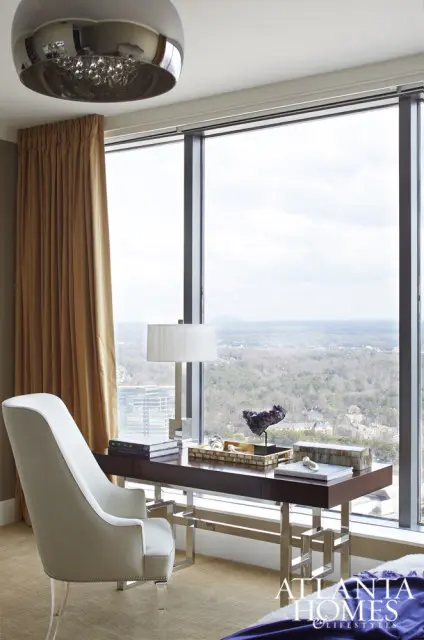 Cream upholstered armchair with acrylic legs next to a dark wood desk with a lamp, books, and amethyst geode, overlooking a city view through a large window with gold curtains.