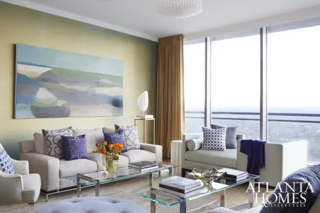Living room with neutral sofas, glass coffee tables, abstract wall art, and large windows overlooking a city skyline.