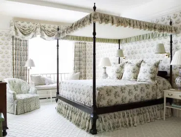 Bedroom with a dark wood four-poster bed, floral print bedding, and matching wallpaper. Features a tufted armchair, a window seat, and a dark wood dresser.