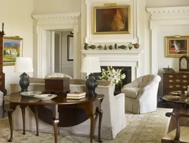 Living room with a tall wooden chest, fireplace with artwork, upholstered chairs, and a round wooden table with decorative plates.